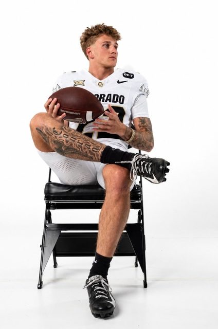 A tattooed athletic jock in a white football uniform sits on a chair holding a football against a white background.