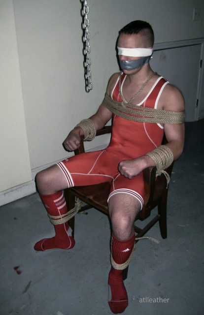 A submissive man in a red wrestling singlet and socks is blindfolded, gagged with duct tape, and tied to a wooden chair with rope.