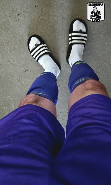 A point-of-view shot of a man with hairy legs wearing purple gym shorts, blue compression sleeves, white tube socks, and striped slides on a concrete floor.