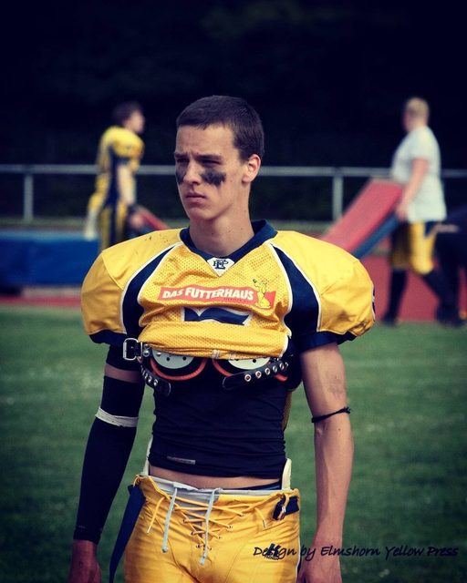 A young football player wearing a yellow jersey rolled up over shoulder pads stands on an outdoor field.
