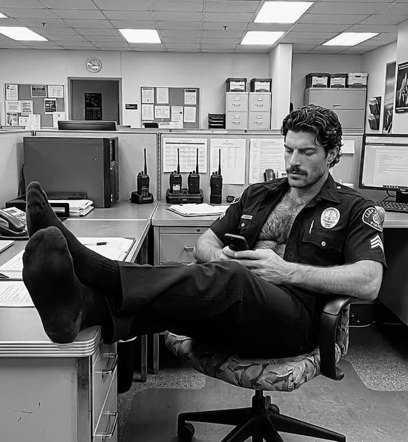 A muscular man in an unbuttoned police uniform sits with his feet up on a desk in an office.