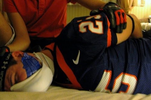 A man in an American football jersey and gloves lies bound and gagged with blue tape while being pinned down.