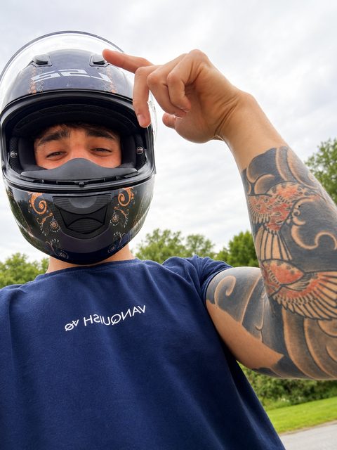 A muscular, tattooed man wears a full-face motorcycle helmet and a navy blue t-shirt while standing outdoors.