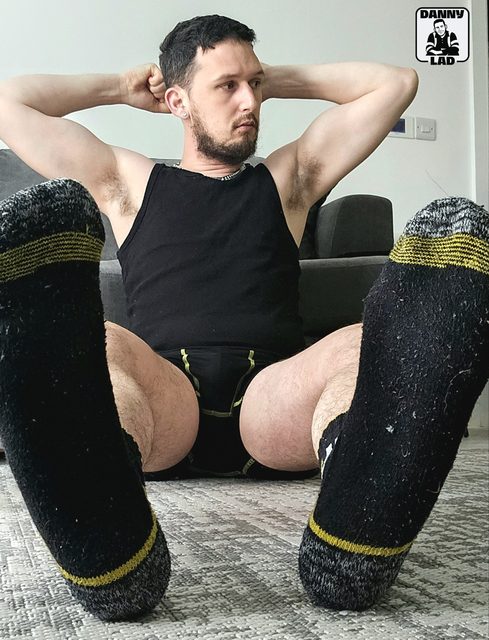 A bearded man sits on the floor wearing a black tank top, briefs, and thick socks with his feet angled toward the camera.
