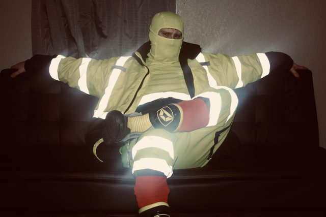 A man sits on a dark couch wearing a full high-visibility workwear uniform and a matching yellow balaclava.