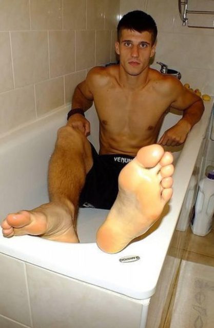 A shirtless muscular twunk sits in a bathtub wearing black athletic shorts, prominently displaying his bare feet and soles toward the camera.