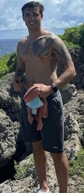 A shirtless, tattooed man wearing gym shorts stands on a rocky coastline with the ocean in the background.