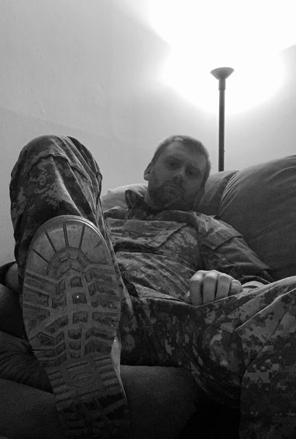A bearded man in a full digital camo military uniform reclines on a sofa, presenting the sole of his combat boot toward the camera.