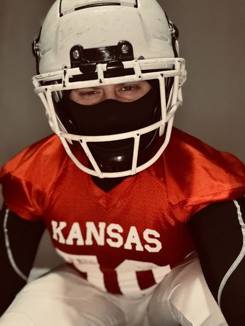 A man wearing a full red Kansas football uniform, shoulder pads, and a white helmet poses while wearing a black balaclava.