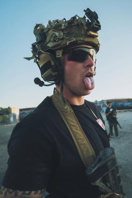 A fit man wearing tactical military gear including a helmet and communication headset sticks his tongue out while outdoors.