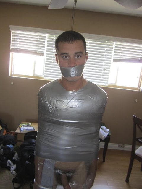 A man is bound around his torso and mouth with silver duct tape, with plastic wrap covering his lower body in a home setting.