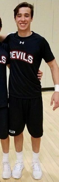 A smiling young man stands in a gymnasium wearing a black athletic t-shirt, black gym shorts, white crew socks, and white sneakers.