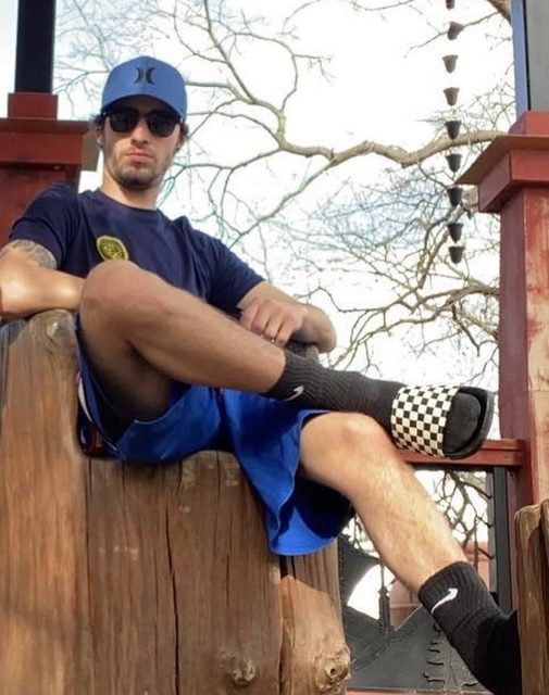 A bearded man in gym shorts and a baseball cap sits outdoors wearing black crew socks with checkered slides.