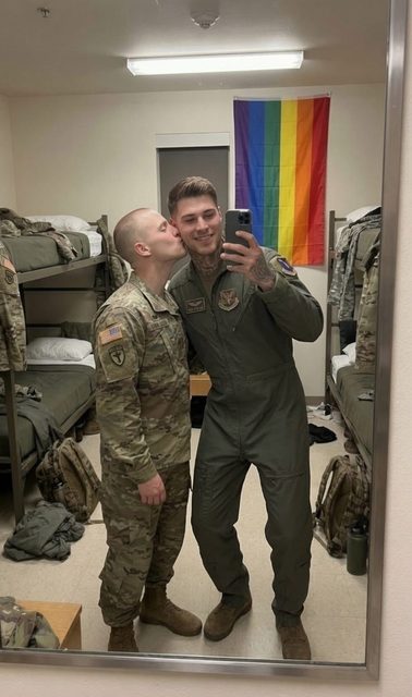 Two men in military uniforms, one in camo fatigues and the other in a green flight suit, share a kiss in a barracks room featuring a pride flag.