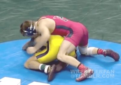 Two male wrestlers in singlets and headgear grapple on a wrestling mat during a match.