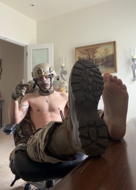 A muscular man in tactical gear and a mask relaxes with one boot and one bare foot on a table.