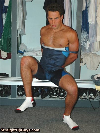 A muscular jock sits on a locker room bench wearing a wrestling singlet pulled down to his waist with his arms behind his back.