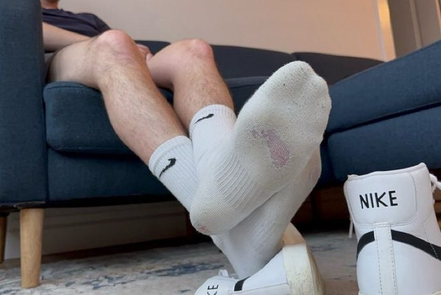 A man with hairy legs relaxes on a sofa, displaying the soles of his white Nike crew socks with his sneakers nearby.