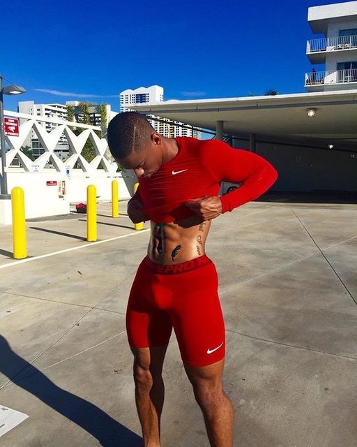 A muscular young man in red compression gear poses outdoors while lifting his shirt to showcase his abdominal muscles and tattoos.