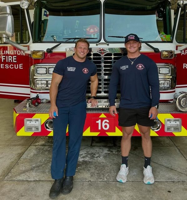 Two muscular men in firefighter uniforms pose together in front of a fire engine at a station.