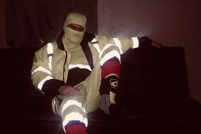 A mature man sits on a dark couch wearing a full hi-vis reflective suit and a matching balaclava.