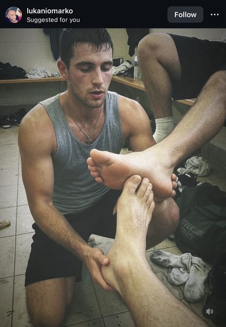 A muscular jock in a grey tank top kneels in a gym locker room to worship and massage another man's sweaty feet.