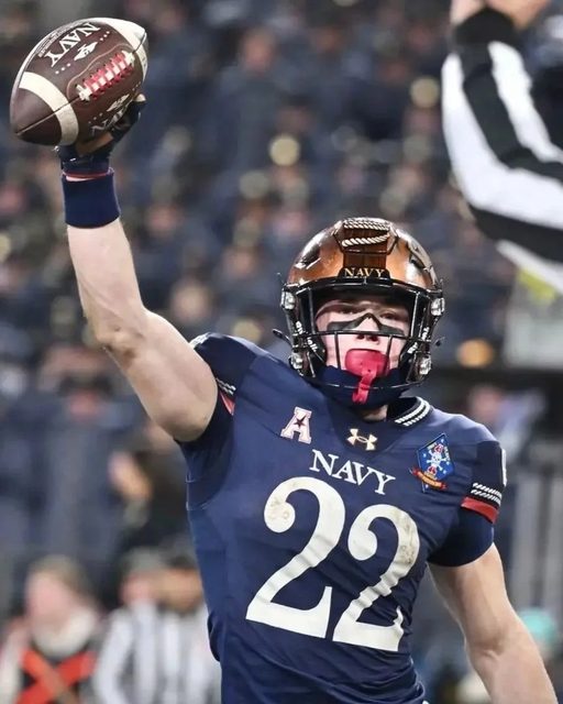 A muscular football player in full Navy uniform holds a ball aloft while wearing his helmet and a red mouthguard.