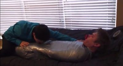 A man leans over a submissive partner who is wrapped tightly in silver duct tape and gagged on a bed.