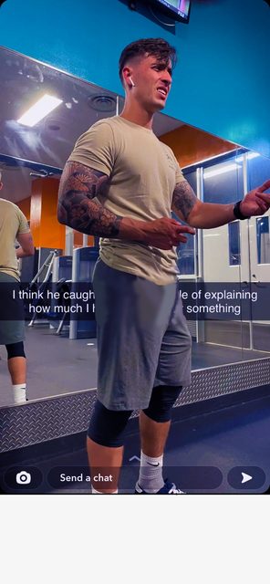A muscular, tattooed man wearing a t-shirt and grey gym shorts over compression sleeves stands in a gym, showing a prominent bulge.