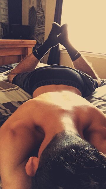A shirtless man lies face down on a bed with his legs bent and socked feet crossed in the air.