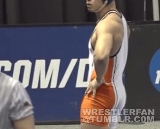 A male wrestler with an athletic body type stands wearing an orange and white wrestling singlet.