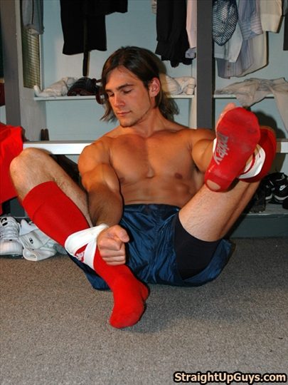 A muscular shirtless young man sits on the locker room floor wearing blue athletic shorts and bright red knee-high socks.
