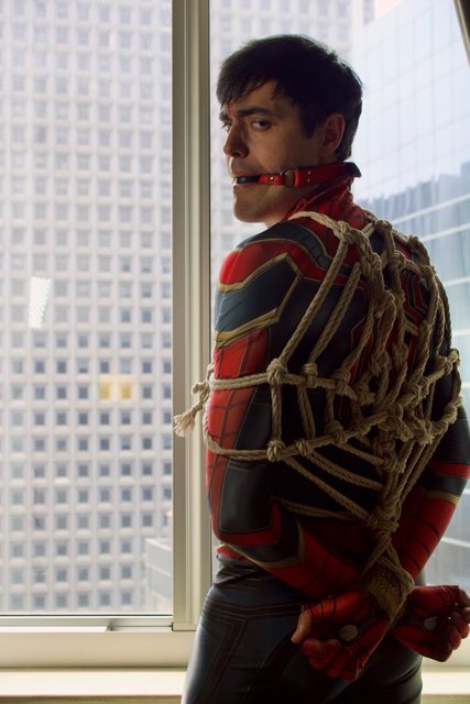 A man wearing a spandex cosplay suit is bound with intricate rope bondage and gagged with a red ball gag in front of a high-rise window.