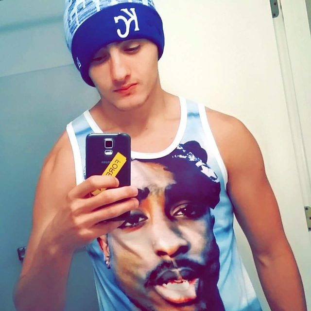 A young man wearing a beanie and a graphic tank top takes a mirror selfie indoors.
