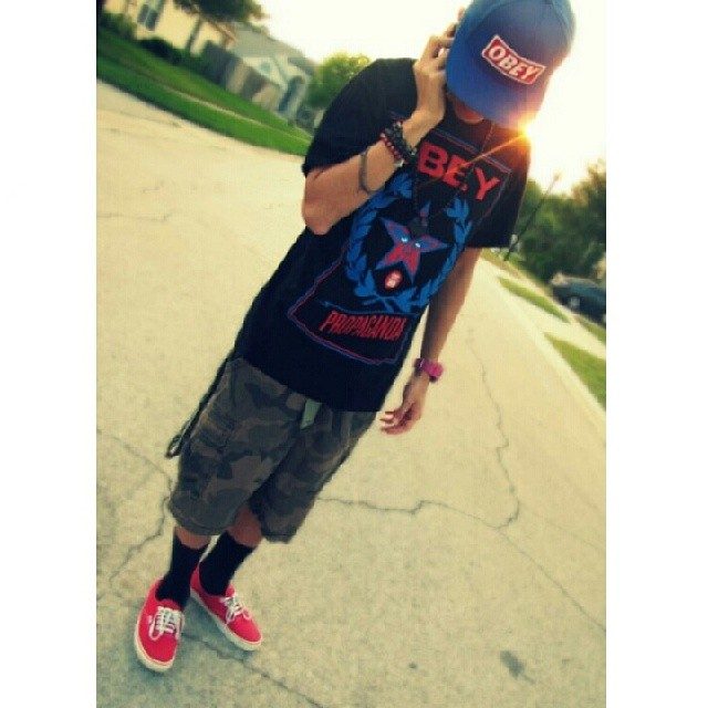 A young man in streetwear, featuring a blue cap, graphic tee, camo shorts, and red sneakers, stands on a residential street.