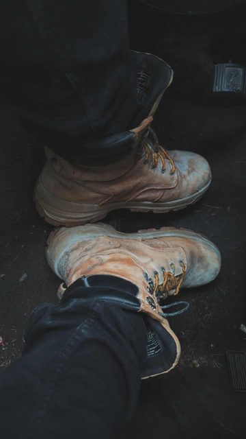 A pair of dirty tan work boots are worn with dark denim jeans in the footwell of a vehicle.