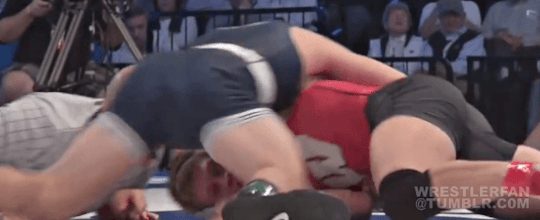Two young men in wrestling singlets engage in a grappling match on an indoor mat.