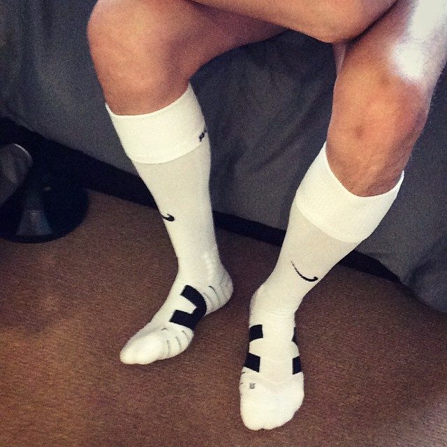 A man's legs and feet are shown wearing white athletic socks, sitting on a bed with his feet on the floor.
