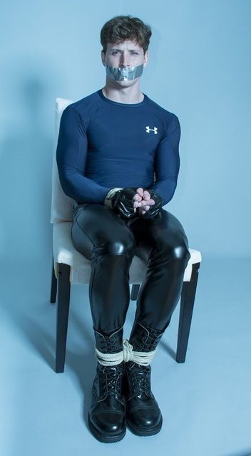 A young athletic man in a blue compression shirt and black leather-look pants is bound to a chair with rope and gagged with duct tape.
