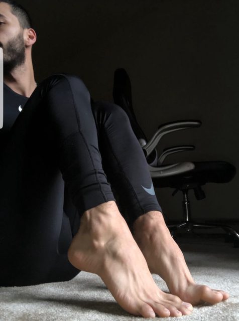 A bearded man wearing black spandex compression gear sits on a carpeted floor with his bare feet prominently displayed in the foreground.