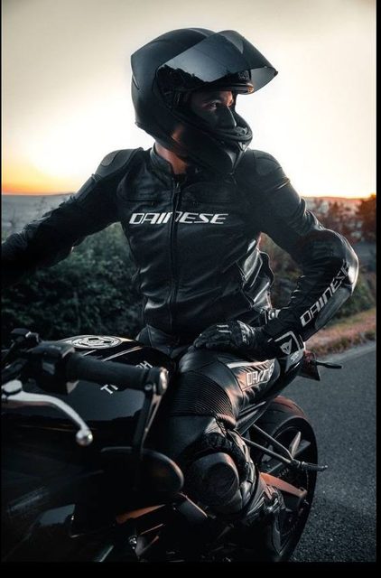A rider dressed in a full black leather motorcycle suit and helmet poses on a sports bike outdoors at sunset.