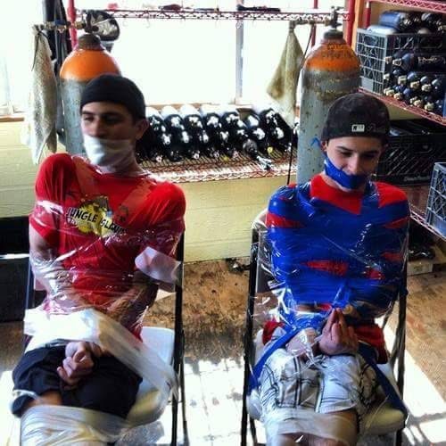 Two young men sit bound to chairs with plastic wrap and duct tape inside a workshop filled with gas tanks.