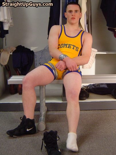 A muscular young man in a yellow wrestling singlet sits in a locker room with his wrists and thighs bound in silver duct tape.
