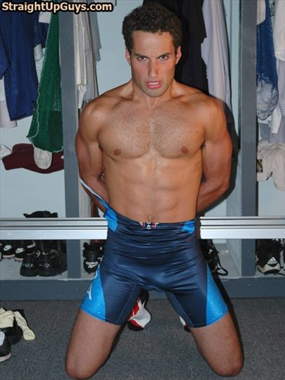 A muscular jock kneels in a locker room wearing a blue wrestling singlet peeled down to his waist with his hands behind his back.