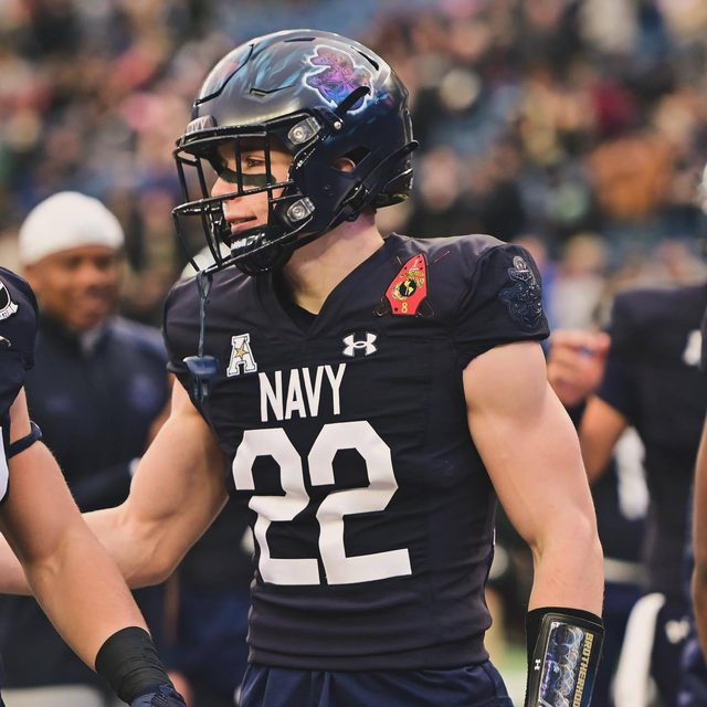 A muscular young athlete wears a tight navy blue football uniform and helmet on a field.