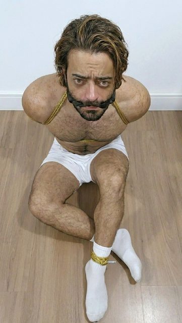 A hairy, bearded man sits on a wooden floor, bound with gold rope around his chest and ankles while wearing a black gag.