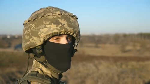 A young man wears a full digital camouflage military uniform with a helmet and black balaclava while standing outdoors.