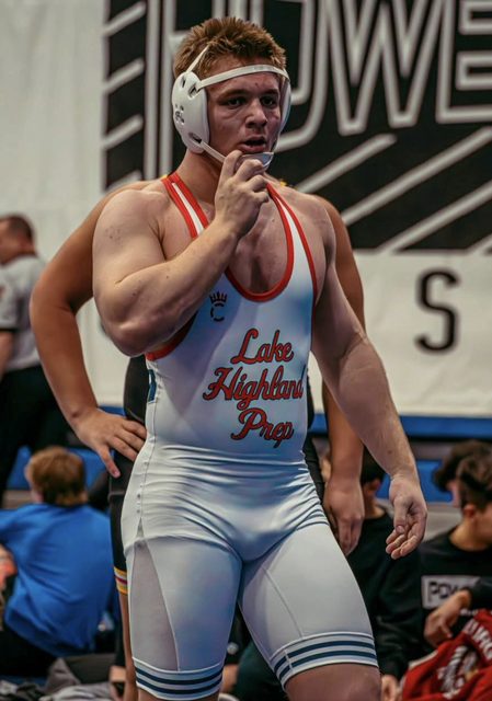 A muscular wrestler stands in a gym wearing a white Lake Highland Prep singlet and headgear.