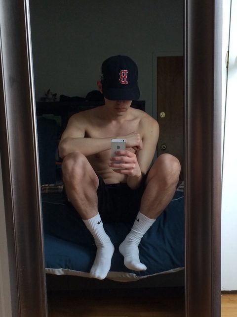 A shirtless young man wearing a baseball cap and dark shorts sits on a bed taking a mirror selfie while wearing white crew socks.