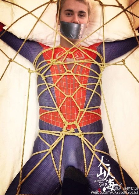 A young man in a Spiderman spandex suit is gagged with duct tape and intricately bound with ropes on a bed.